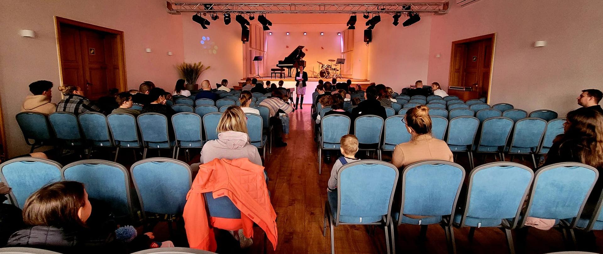 Sala koncertowa PSM w Gostyniu podczas inauguracji dni otwartych szkoły. Na scenie stoi fortepian oraz perkusja. Zgromadzeni słuchacze słuchają wypowiedzi Pani Dyrektor Katarzyny Grzybowskiej. 