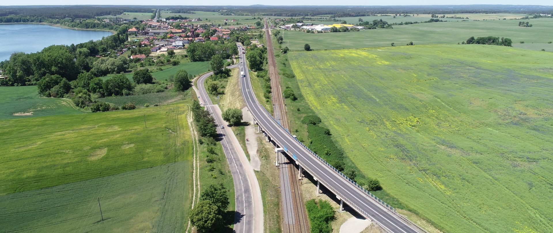 Zdjęcie lotnicze przedstawia rozległy, zielony krajobraz z polami uprawnymi i niewielką miejscowością po lewej stronie, położoną nad dużym jeziorem. Przez środek kadru biegnie równolegle droga oraz linia kolejowa, która miejscami przechodzi nad terenem na estakadzie. Po prawej stronie widoczne są rozciągające się pola w różnych odcieniach zieleni, a horyzont wypełnia pas lasu.