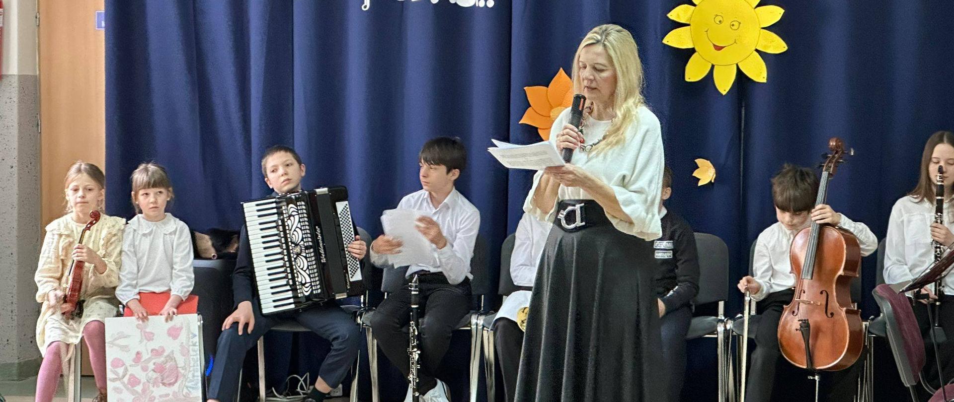 Anna Łysko prowadzi audycje dla uczniów. W tle siedzą wykonawcy - sześcioro uczniów wraz z instrumentami.