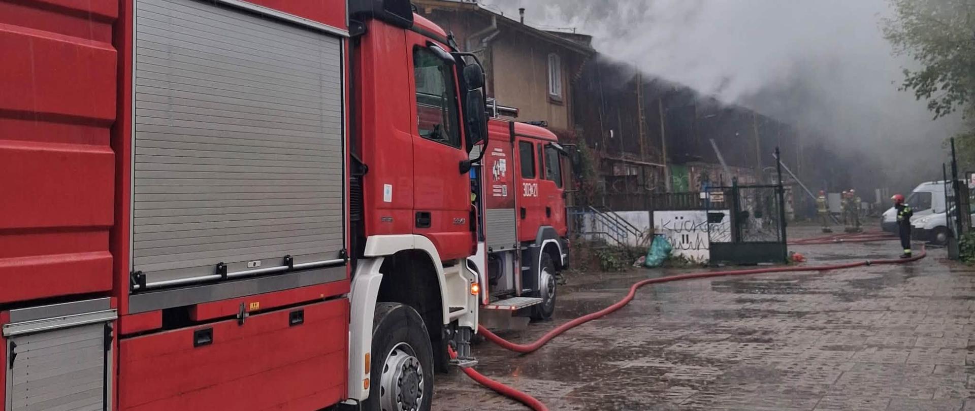 Perspektywa boczna z poziomu ulicy, pokazująca kilka wozów strażackich zaparkowanych jeden za drugim. Czerwone pojazdy, wyposażone w drabiny i sprzęt, zajmują większą część kadru po lewej stronie. Na mokrej nawierzchni rozciągnięte są czerwone węże strażackie. W oddali widać płonący budynek i kłęby dymu, a przed nim kilku strażaków walczących z ogniem. W tle, po prawej stronie, widać zaparkowane samochody osobowe.