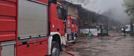 Perspektywa boczna z poziomu ulicy, pokazująca kilka wozów strażackich zaparkowanych jeden za drugim. Czerwone pojazdy, wyposażone w drabiny i sprzęt, zajmują większą część kadru po lewej stronie. Na mokrej nawierzchni rozciągnięte są czerwone węże strażackie. W oddali widać płonący budynek i kłęby dymu, a przed nim kilku strażaków walczących z ogniem. W tle, po prawej stronie, widać zaparkowane samochody osobowe.