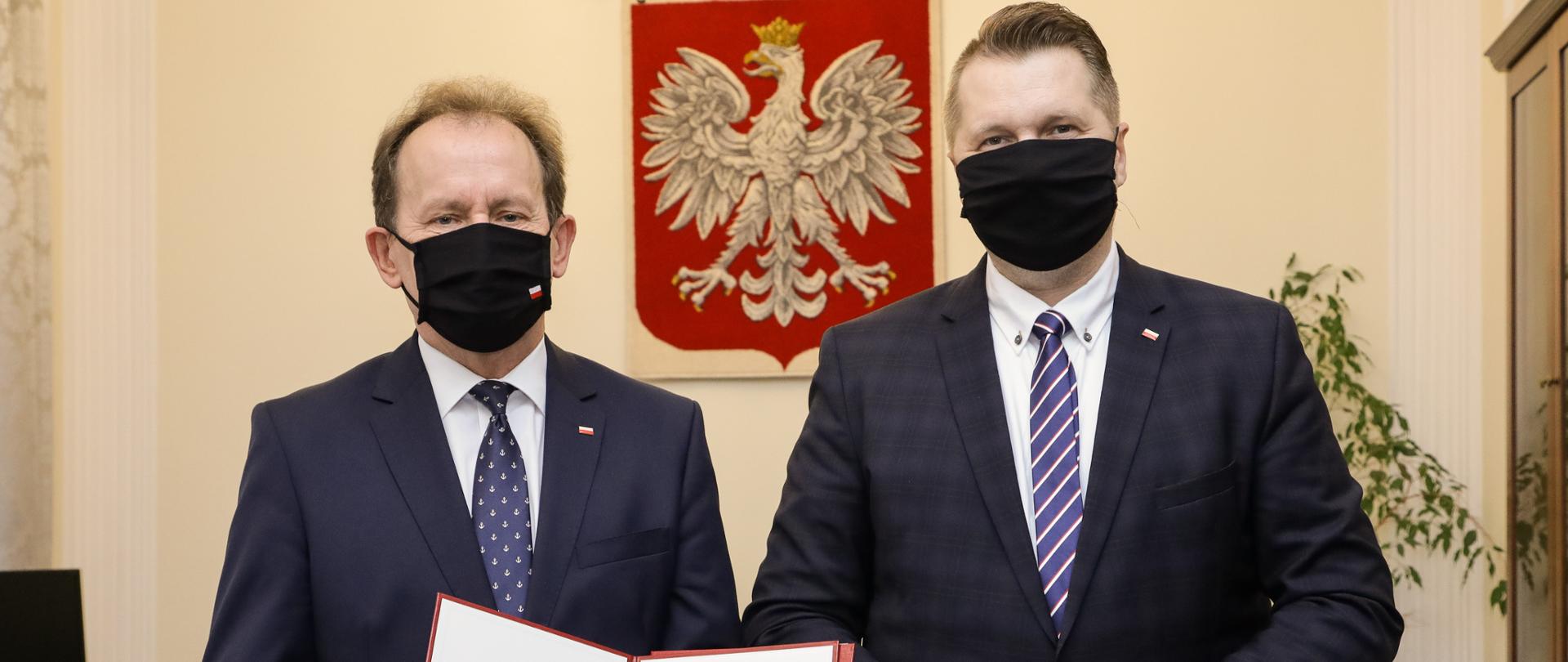 Prof. Bernacki i minister Czarnek stoją obok siebie, , prof. Bernacki trzyma nominację, za nimi na ścianie godło Polski.