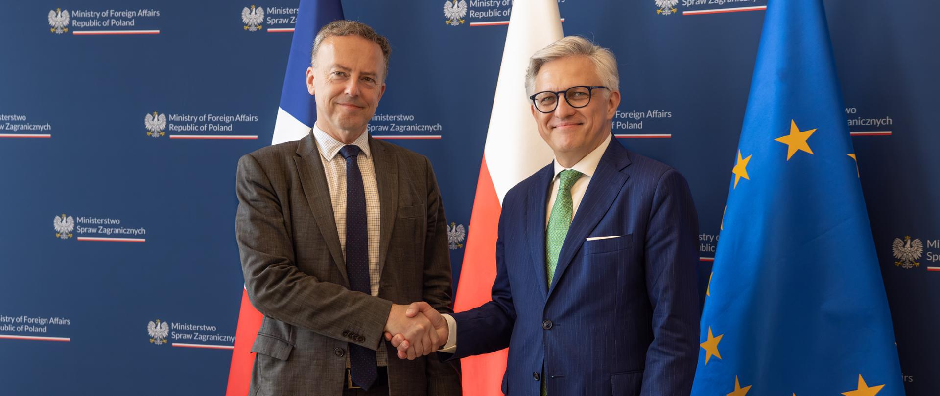 Martin Briens, Secretary General of the French Ministry of Europe and Foreign Affairs, and Artur Harazim, Undersecretary of State, shake hands against a navy blue backdrop featuring the logos of the Polish Ministry of Foreign Affairs and the flags of France, Poland, and the European Union. Both are smiling for the camera.