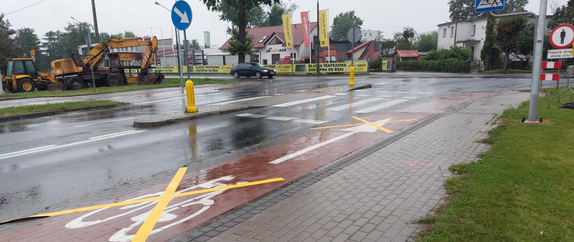 Na zdjęciu widać mokrą ulicę z różnymi znakami drogowymi i oznaczeniami. Jest tam ścieżka rowerowa oznaczona żółtymi krzyżykami, co wskazuje, że jest nieczynna. Po prawej stronie zdjęcia znajduje się znak przejścia dla pieszych. Po lewej stronie ulicy zaparkowane są pojazdy budowlane, a w tle widać budynki. Nawierzchnia jest mokra, co sugeruje, że niedawno padał deszcz.