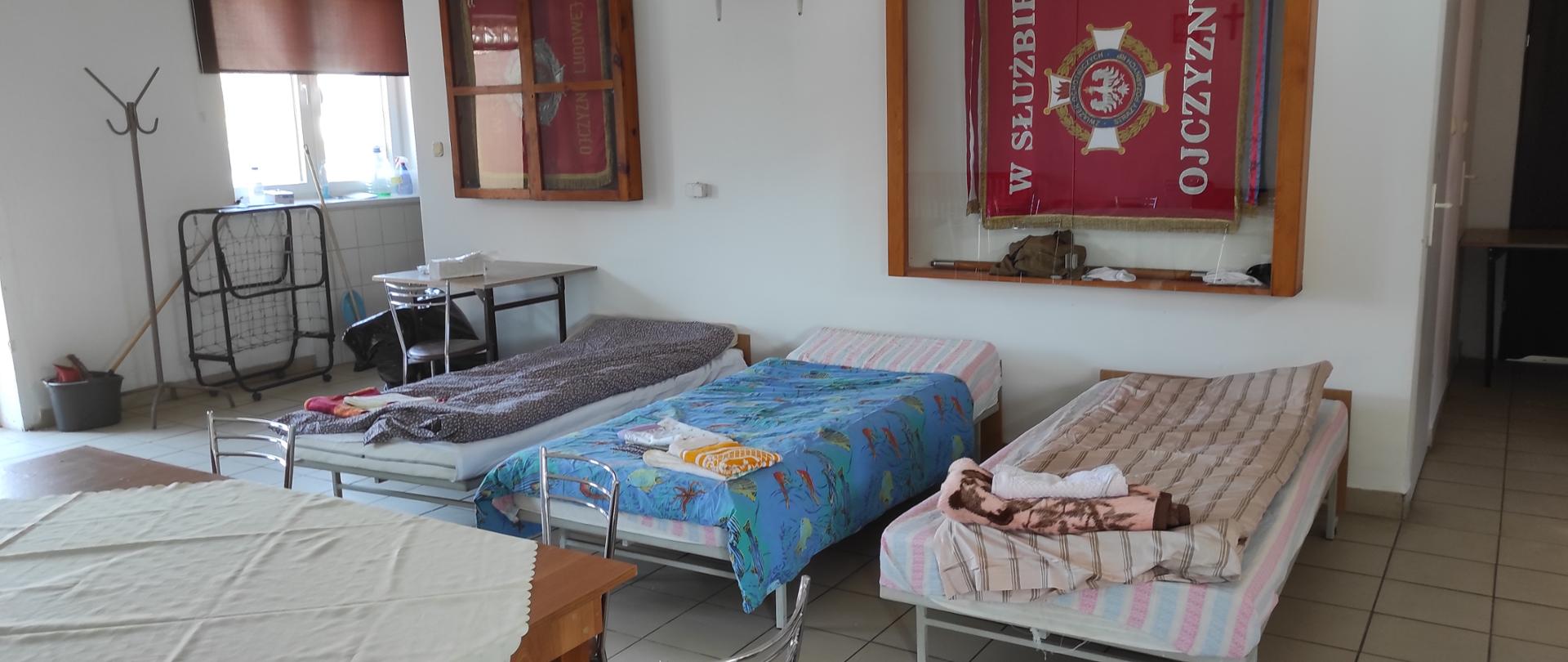 Świetlica OSP. na pierwszym planie znajdują się trzy posłane łóżka. Na ścianie dwie gabloty ze sztandarami OSP. Z lewej strony stolik przykryty jasnym obrusem.