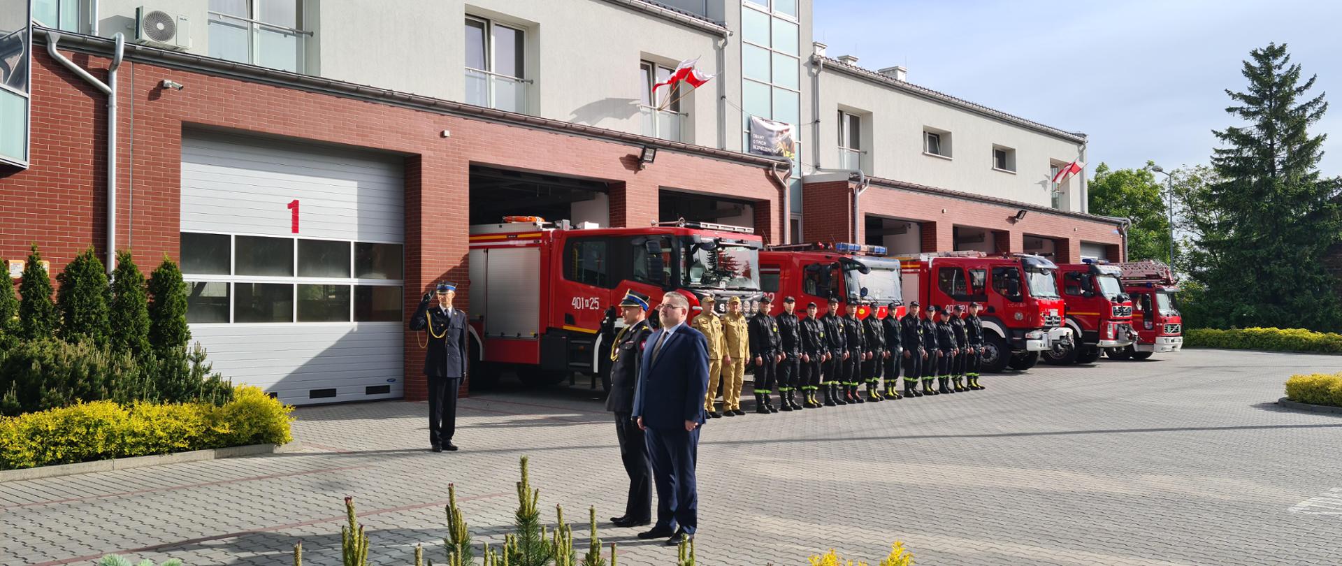 Zdjęcie przedstawia strażaków, którzy stoją na placu przed garażami oraz starostę powiatu bocheńskiego. Część strażaków ubrana jest w stroje galowe, a część w stroje bojowe. Komendant Powiatowy PSP w Bochni salutuje z okazji podniesienia flagi państwowej