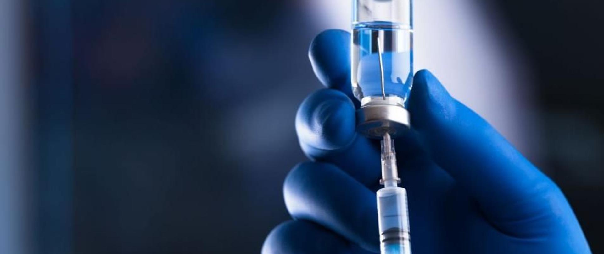 Na zdjęciu znajduje się strzykawka wraz z fiolką szczepionki trzymane w dłoni medyka. Tło jest w odcieniach granatowego.