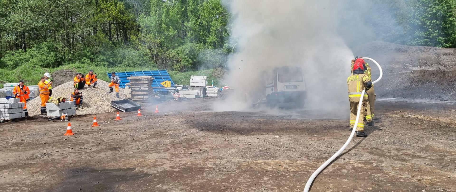 Zdjęcie przedstawia pokaz ratownictwa technicznego przez strażaków. Strażacy gaszą srebrny samochód. Dookoła jest publiczność. 