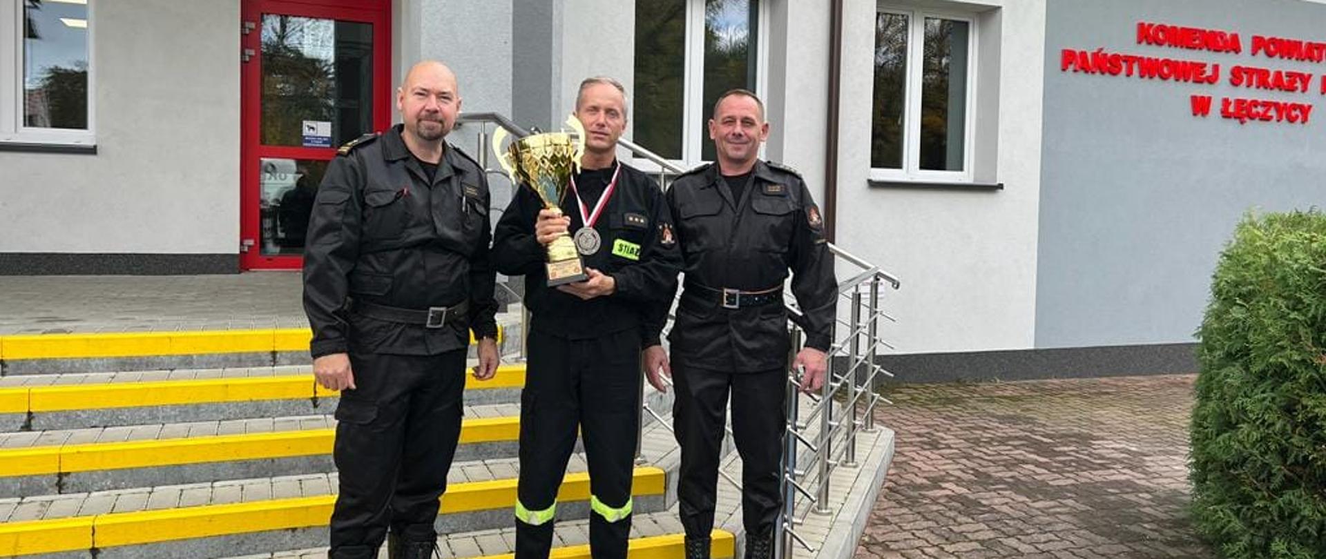 Na zdjęciu widać trzech strażaków ubranych w czarne stroje bez czapek. Strażak w środku trzyma słoty puchar a na szyi zawieszony ma medal. Strażacy stoją na schodach. W tle widać biało-szary budynek Komendy Powiatowej Państwowej Straży Pożarnej w Łęczycy. Jest widno.