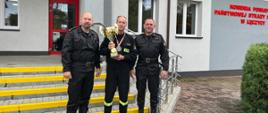 Na zdjęciu widać trzech strażaków ubranych w czarne stroje bez czapek. Strażak w środku trzyma słoty puchar a na szyi zawieszony ma medal. Strażacy stoją na schodach. W tle widać biało-szary budynek Komendy Powiatowej Państwowej Straży Pożarnej w Łęczycy. Jest widno.