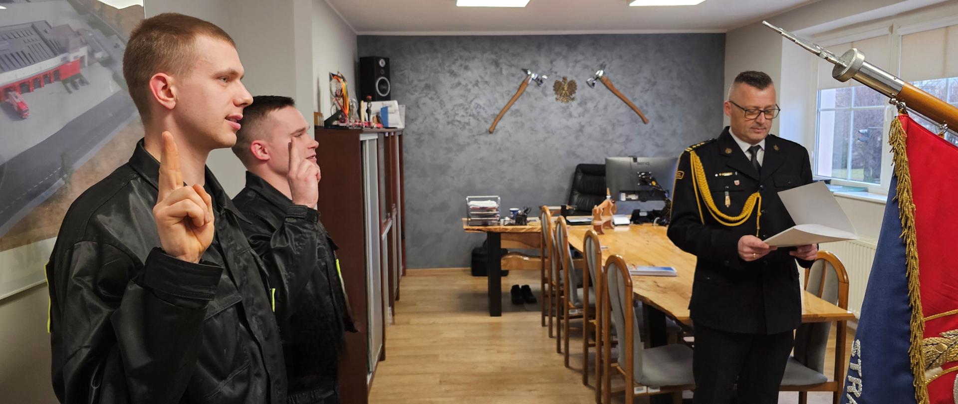 Gabinet, w którym zbiórkę maja strażacy. Dwóch stoi po lewej stronie i z podniesionymi prawymi rękoma składa ślubowanie. Jeden z teczką stoi na wprost za sztandarem.