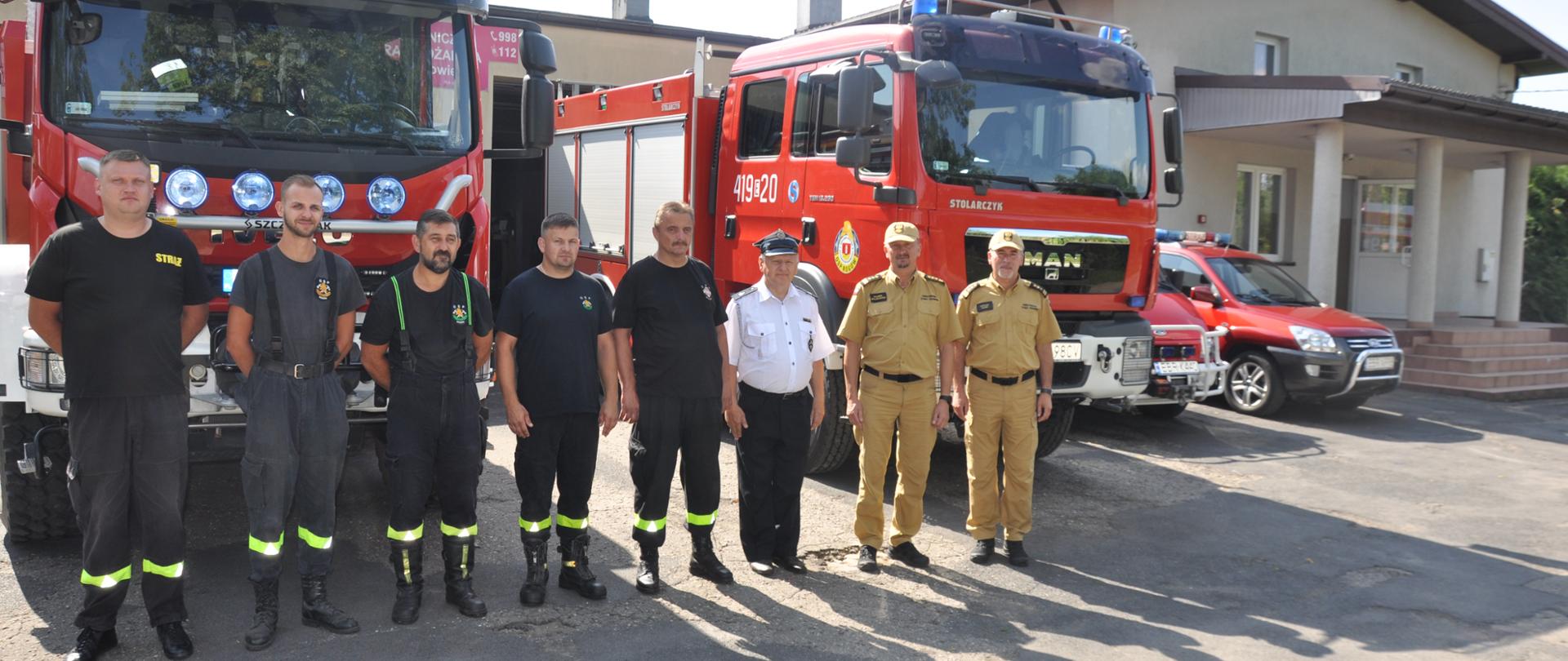 Na pierwszym planie w szeregu sześciu strażaków OSP Rogów, komendant powiatowy PSP w Brzezinach oraz zastępca komendanta wojewódzkiego PSP w Łodzi starszy brygadier Malinowski . W tle za stojącymi w szeregu strażnica OSP Rogów oraz samochody bojowe OSP.