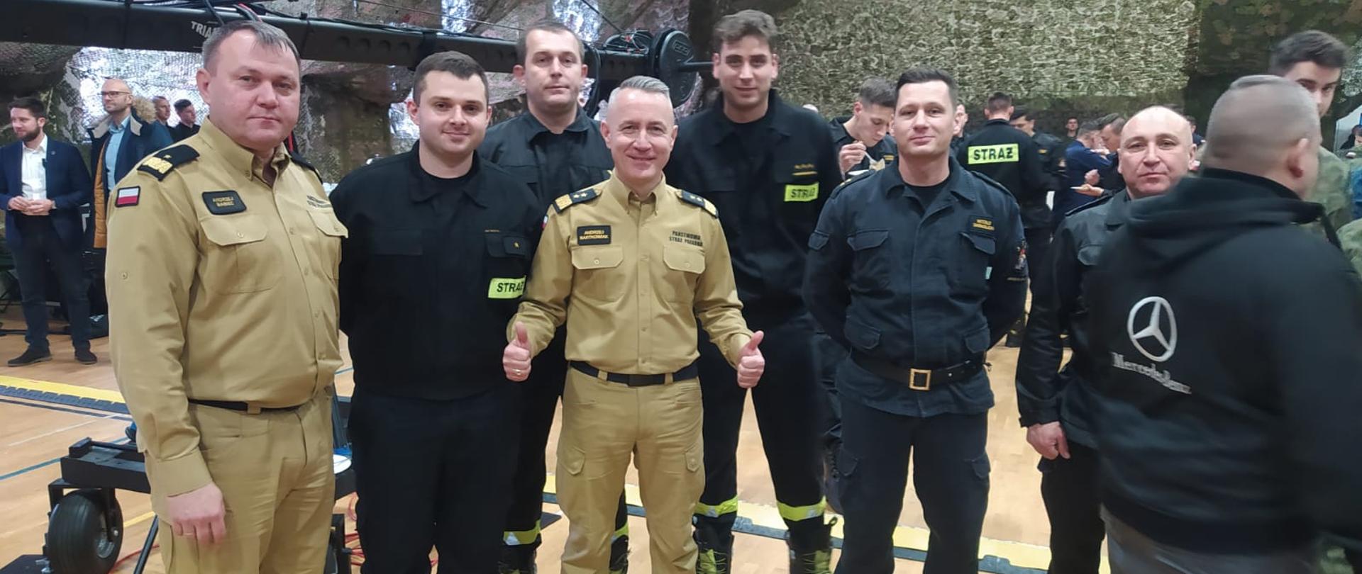 Na zdjęciu widać funkcjonariuszy KP PSP w Ustrzykach Dolnych z Komendantem Głównym PSP oraz Podkarpackim Komendantem Wojewódzkim. Zdjęcie wykonane jest na hali a w tle widać funkcjonariuszy innych służb. 