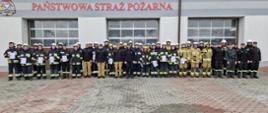 Zdjęcie przedstawia wszystkich uczestników ora Osoby które szkoliły ochotników podczas szkolenia kierowców-konserwatorów sprzętu pożarniczego. Wszyscy stoją w zbiórce w tle widoczne są bramy wjazdowe do garażu JRG.