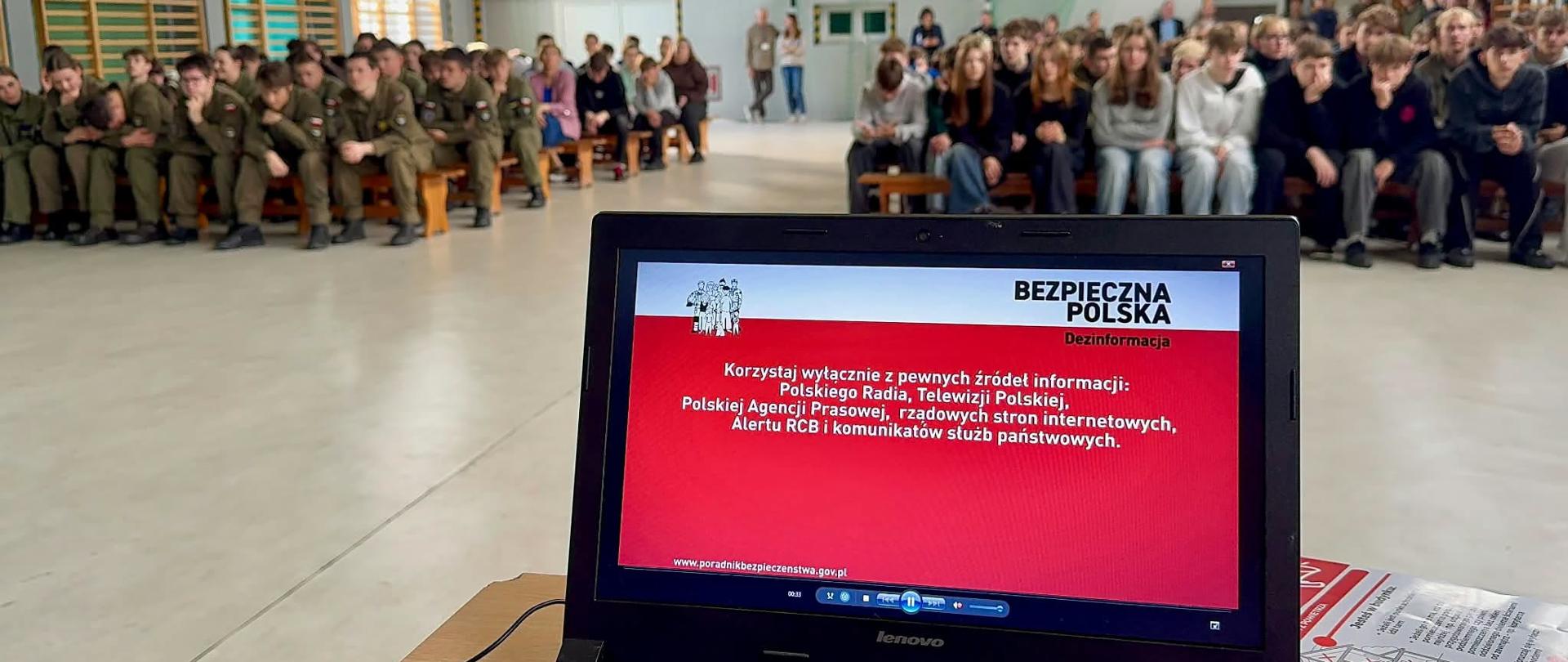 Na zdjęciu widoczna jest sala gimnastyczna, w której zgromadzili się uczniowie oraz młodzież w mundurach. Uczestnicy siedzą na ławkach i słuchają prezentacji poświęconej bezpieczeństwu. Na pierwszym planie znajduje się laptop, na którego ekranie wyświetlany jest materiał edukacyjny „Bezpieczna Polska”, dotyczący dezinformacji i korzystania z wiarygodnych źródeł informacji.
