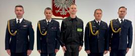 Na zdjęciu widzimy pięciu mężczyzn stojących w szeregu na tle białej ściany z godłem Polski (białym orłem na czerwonym tle) zawieszonym centralnie. Czterech z nich (pierwszy, drugi, czwarty i piąty od lewej) ubranych jest w galowe mundury strażackie. Środkowa osoba (trzecia od lewej) ma na sobie ubranie koszarowe z odblaskowym napisem „STRAŻ” . Zdjęcie wykonane podczas uroczystości ślubowania. Pomieszczenie ma jasną podłogę z paneli i sufit z kasetonami oraz oświetleniem biurowym. Atmosfera formalna i poważna.