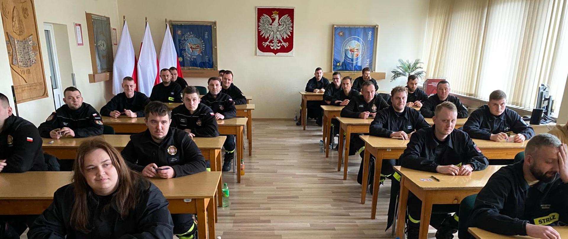 Zdjęcie przedstawia strażaków przed egzaminem teoretycznym w Sali szkoleniowej, w tle widoczne godło Polski.