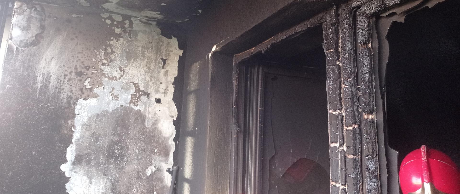 Zdjęcie obrazuje wypalone ściany i futrynę okna oraz hełm strażacki.