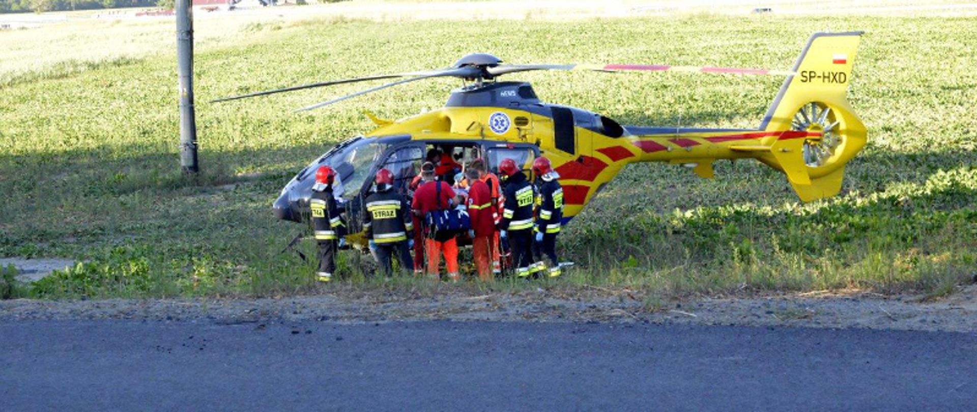 Wypadek Mikoszki. Na polu stoi śmigłowiec Lotniczego Pogotowia Ratunkowego. Strażacy i ratownicy medyczni pomagają w ewakuacji do niego rannego w wypadku chłopca.