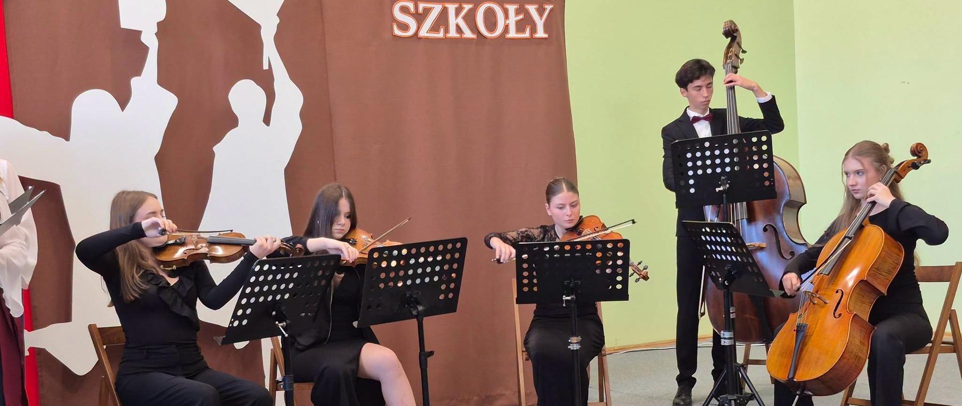Pięcioro uczniów w czarnych strojach gra na skrzypcach, altówce, wiolonczeli i kontrabasie podczas Święta Szkoły w I LO w Mielcu, w tle brązowa kurtyna z napisem „Święto Szkoły".