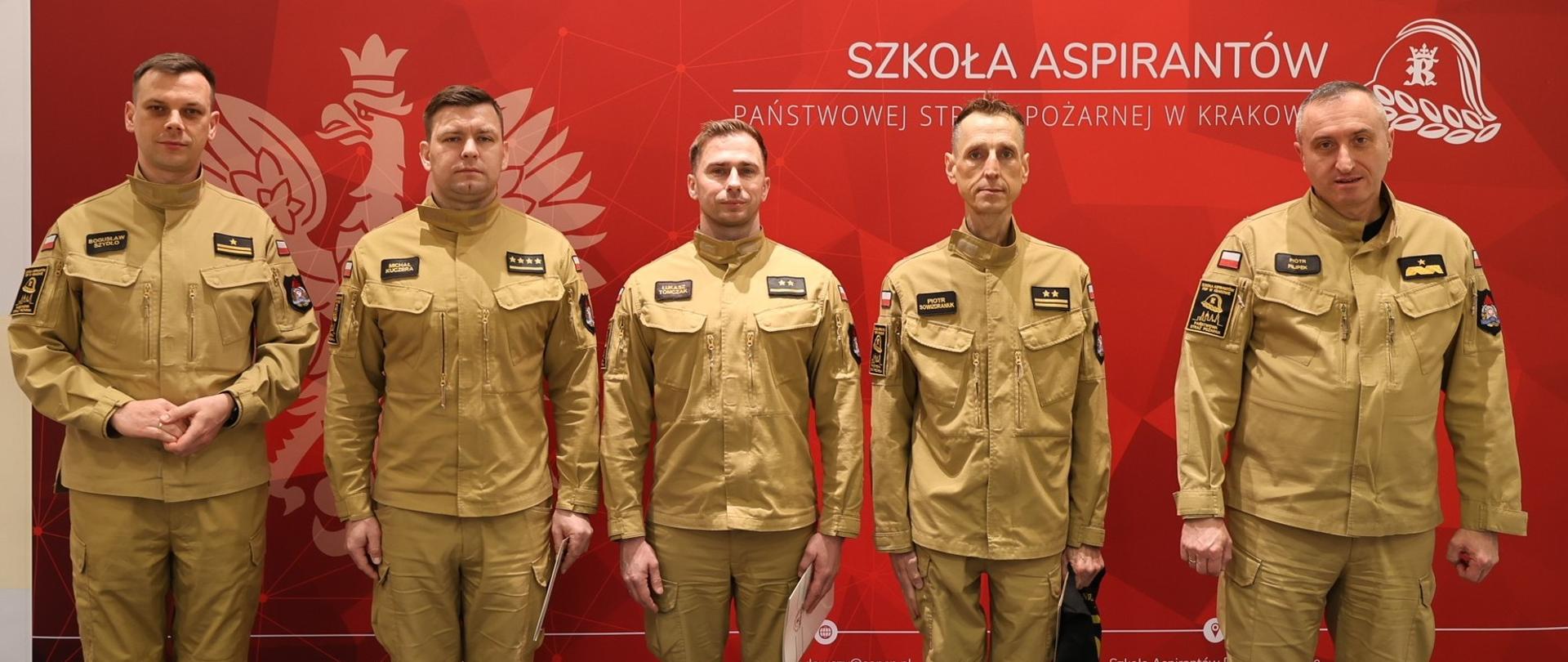 6. Oficjalne zdjęcie pamiątkowe
Pięciu funkcjonariuszy PSP stoi w jednym rzędzie na tle oficjalnej ścianki promocyjnej. Od lewej: Zastępca Komendanta, st. kpt. Michał Kuczera, mł. kpt. Łukasz Tomczak, bryg. Piotr Sowizdraniuk oraz Komendant nadbryg. Piotr Filipek. Wszyscy patrzą prosto w obiektyw.
