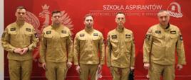 Oficjalne zdjęcie pamiątkowe
Pięciu funkcjonariuszy PSP stoi w jednym rzędzie na tle oficjalnej ścianki promocyjnej. Od lewej: Zastępca Komendanta, st. kpt. Michał Kuczera, mł. kpt. Łukasz Tomczak, bryg. Piotr Sowizdraniuk oraz Komendant nadbryg. Piotr Filipek. Wszyscy patrzą prosto w obiektyw.
