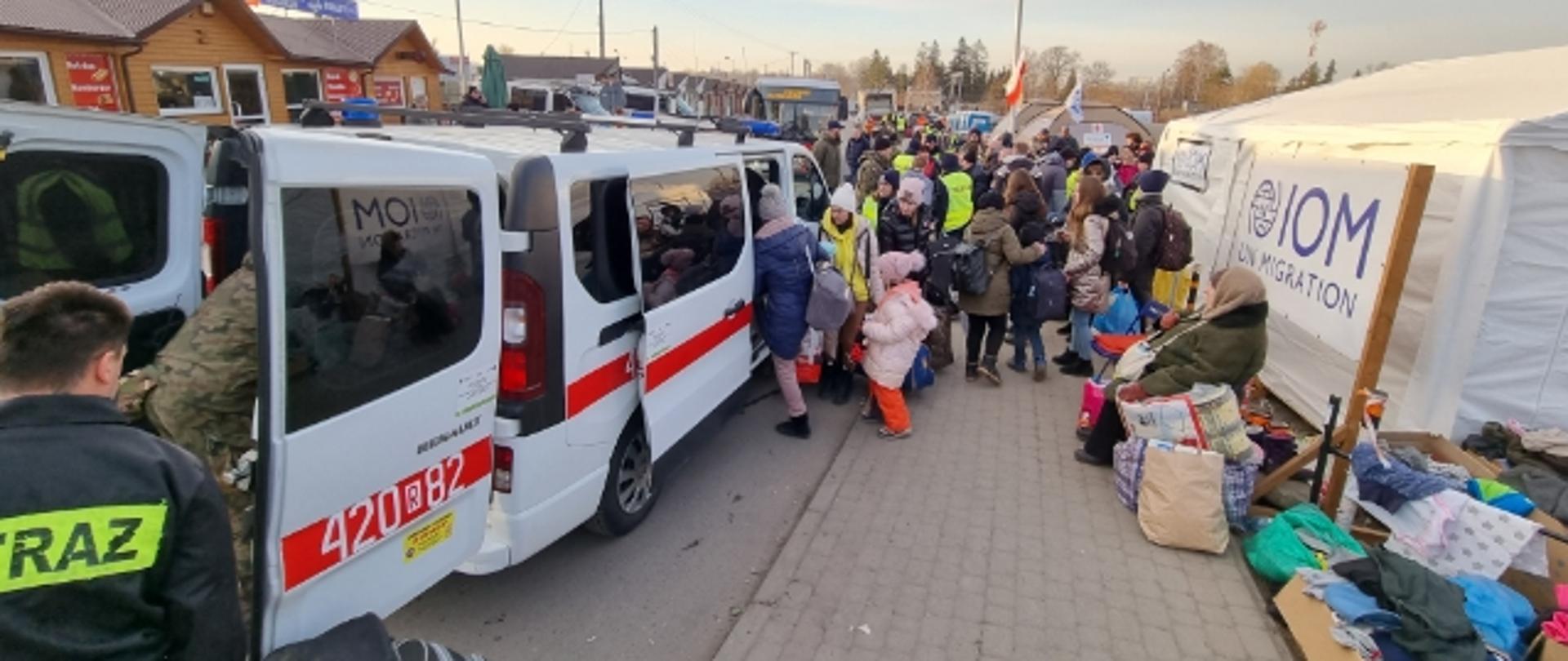 Na pierwszym planie znajduje się bus do którego wsiadają osoby z bagażami. W pobliżu busa jest bardzo dużo osób, w tym strażak Państwowej Straży Pożarnej, który pomaga w załadunku bagaży. Po prawej stronie zdjęcia znajduje się biały namiot. 