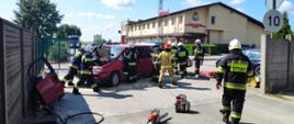 Zdjęcie przedstawia akcję ratowniczą strażaków, którzy prowadzą działania przy samochodzie osobowym. Strażacy odłączają w pojeździe akumulator oraz wykonują dostęp do osób poszkodowanych uwięzionych wewnątrz pojazdu przy wykorzystaniu sprzętu hydraulicznego. Na zdjęciu widać też inny sprzęt: pilarkę do drewna, nosze ortopedyczne oraz linię gaśniczą. W tle zdjęcia widać budynek Komendy Powiatowej PSP w Rypinie i samochód gaśniczy. 