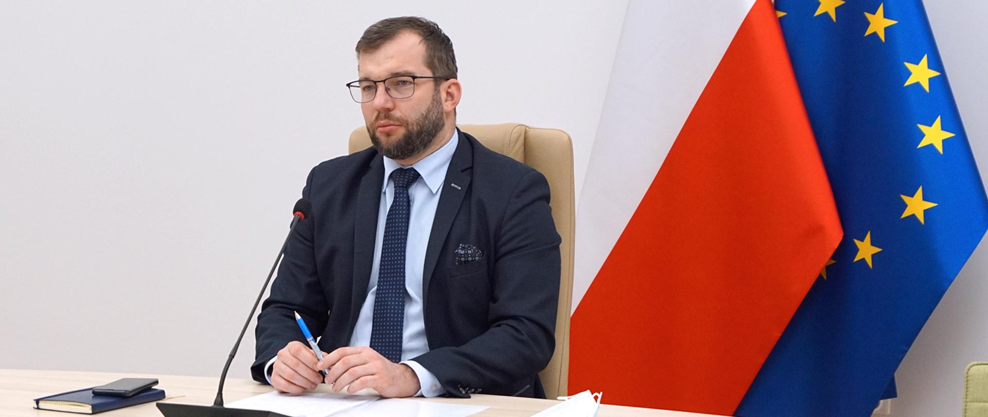 Minister Grzegorz Puda, z długopisem w dłoniach, siedzący na tle flag polskiej i Unii Europejskiej.