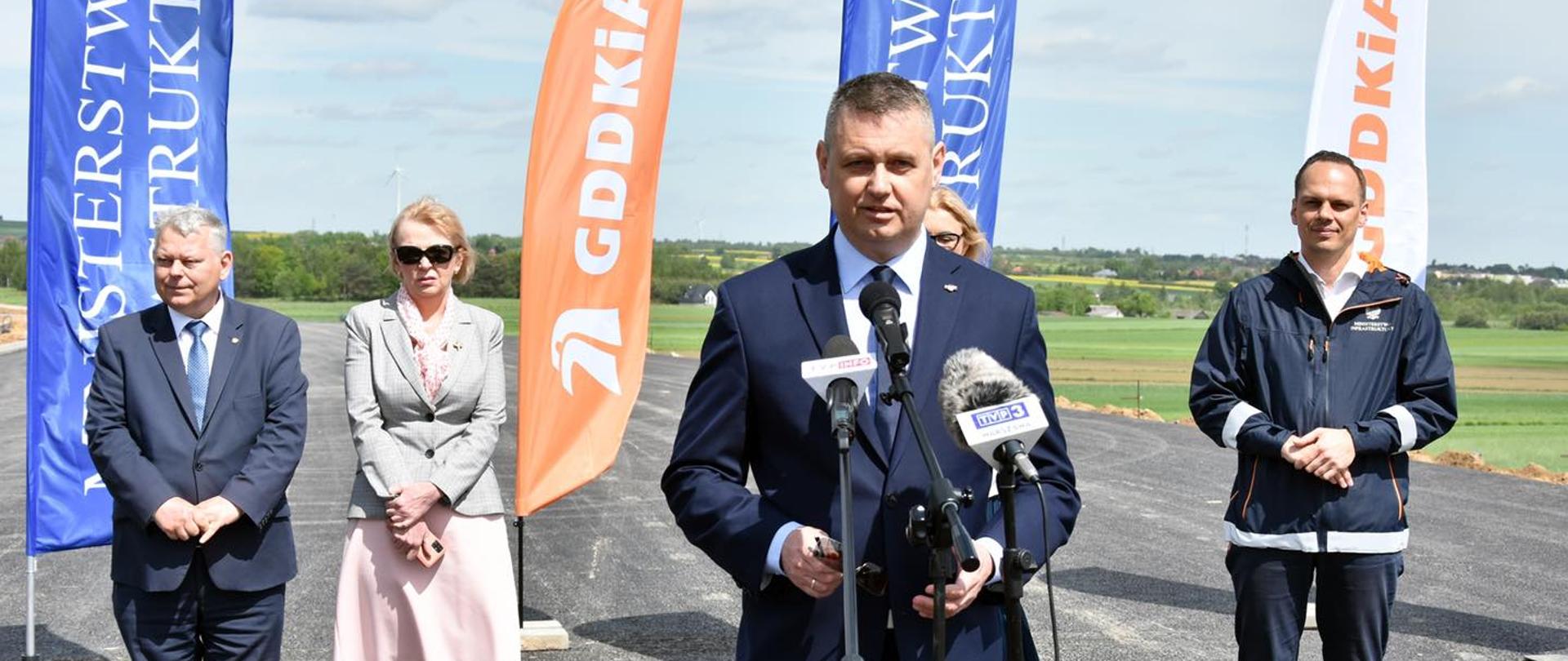 Na zdjęciu na głównym planie znajduje się mężczyzna mówiący do mikrofonu, za nim stoją cztery osoby, wszyscy znajdują się na terenie budowy nowej autostrady.