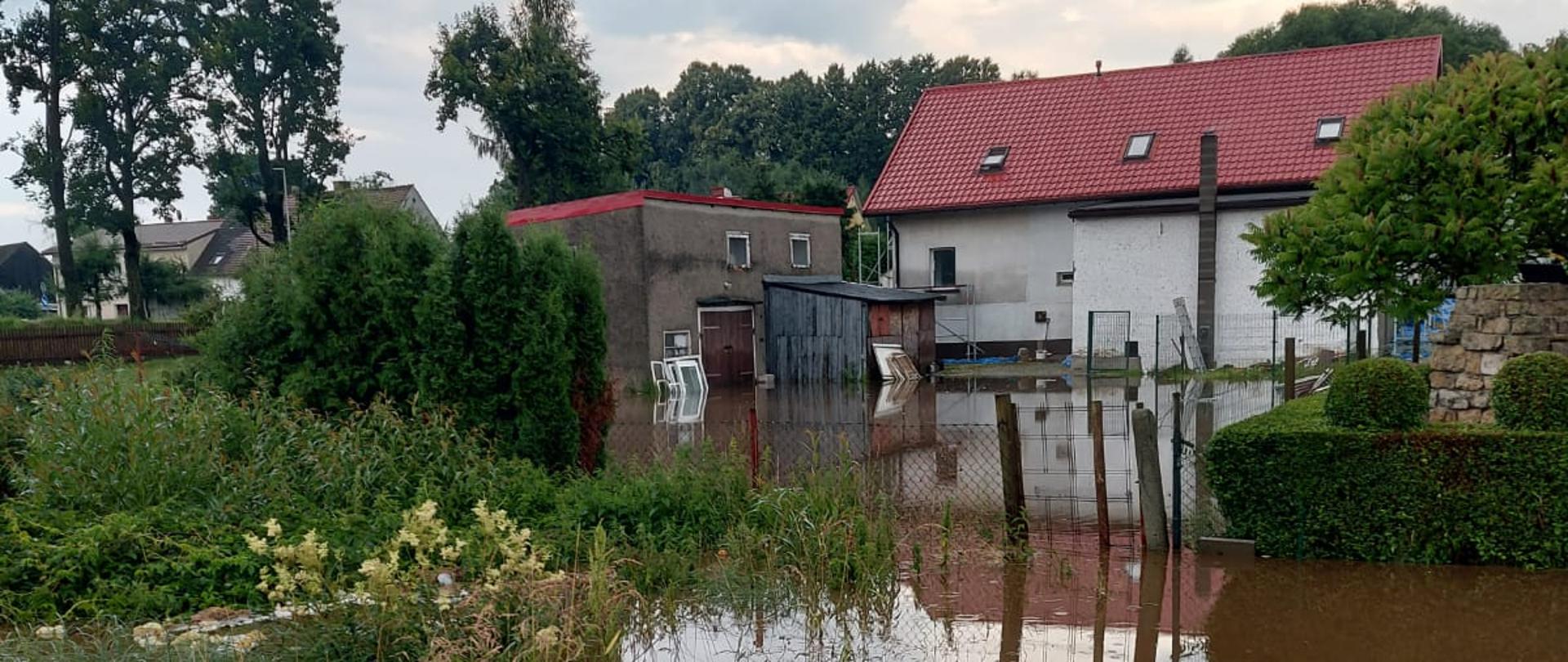 W tle zabudowania mieszkalne w gospodarstwie rolnym. Łąka przed zabudowaniami oraz podwórko zalane wodą, która wezbrała na skutek obfitych opadów deszczu. Z daleka dostrzegamy, że budynek zalany do około 50 cm od poziomu gruntu