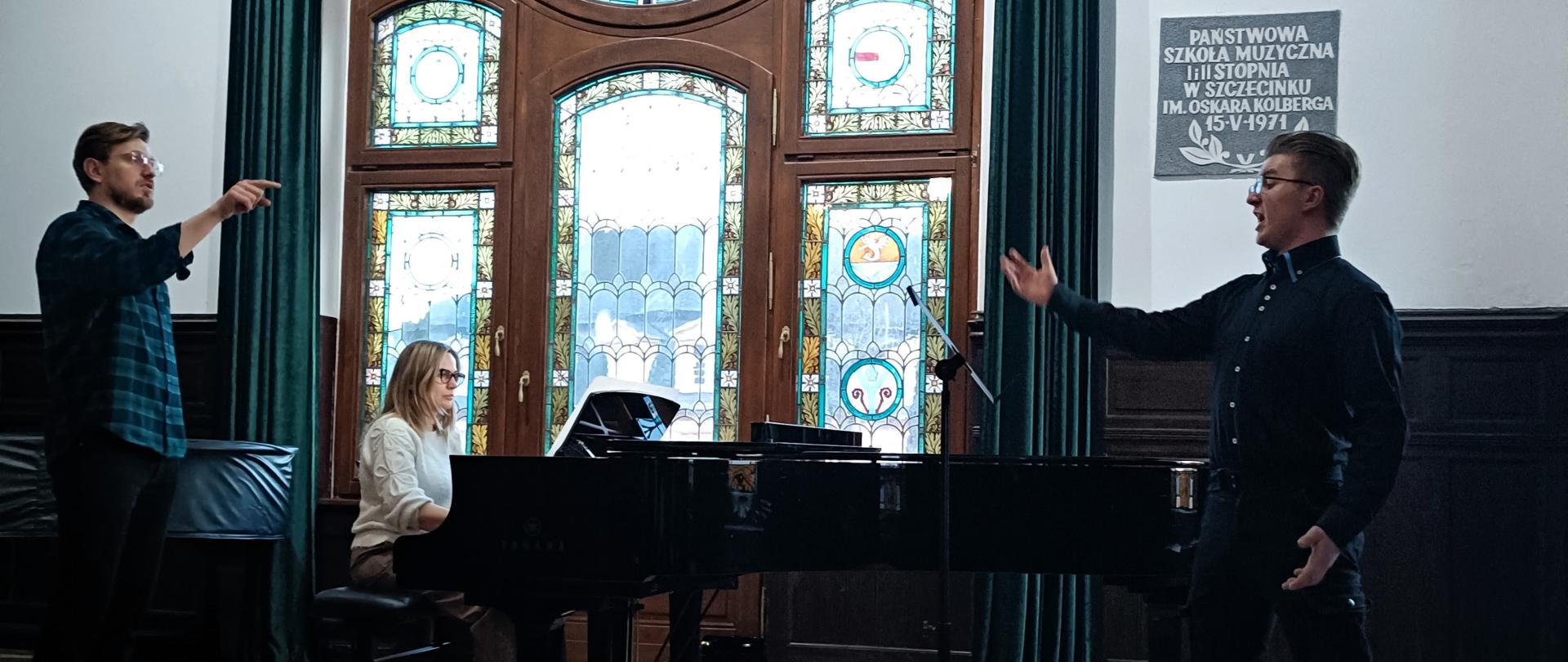 Ujęcie podczas warsztatów wokalnych. Na pierwszym planie stojącego na przeciwko pulpitu ucznia, który śpiewając wyciąga rękę ku górze. Po drugiej stronie sceny stoi instruujący go młody mężczyzna. W tle widać fortepian, przy którym siedzi i gra kobieta w białym swetrze oraz duże witrażowe okno z zielonymi zasłonami.