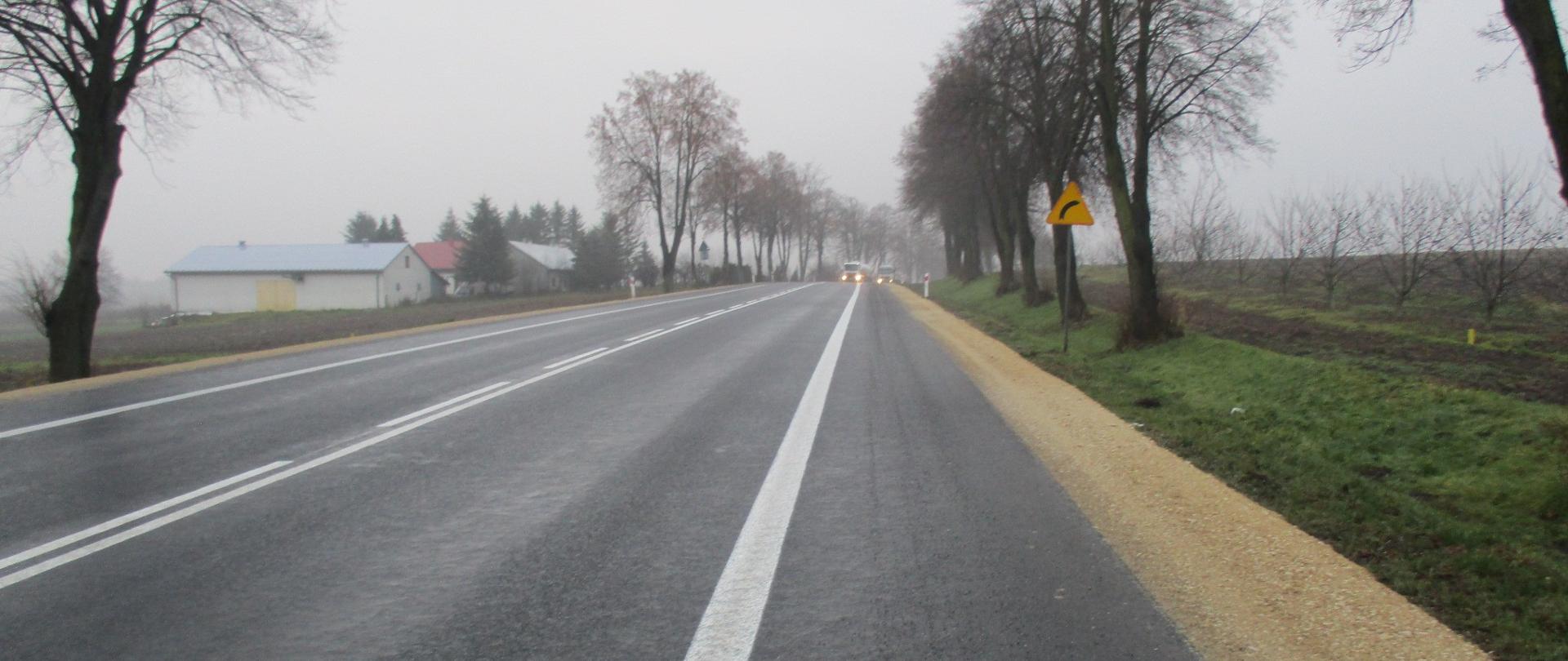 DK78 Słupia - droga jednojezdniowa z namalowanym białym oznakowaniem poziomym, w oddali światła samochodów, przy drodze drzewa, pola i domy, żółty znak drogowy oznaczający zakręt w prawo 