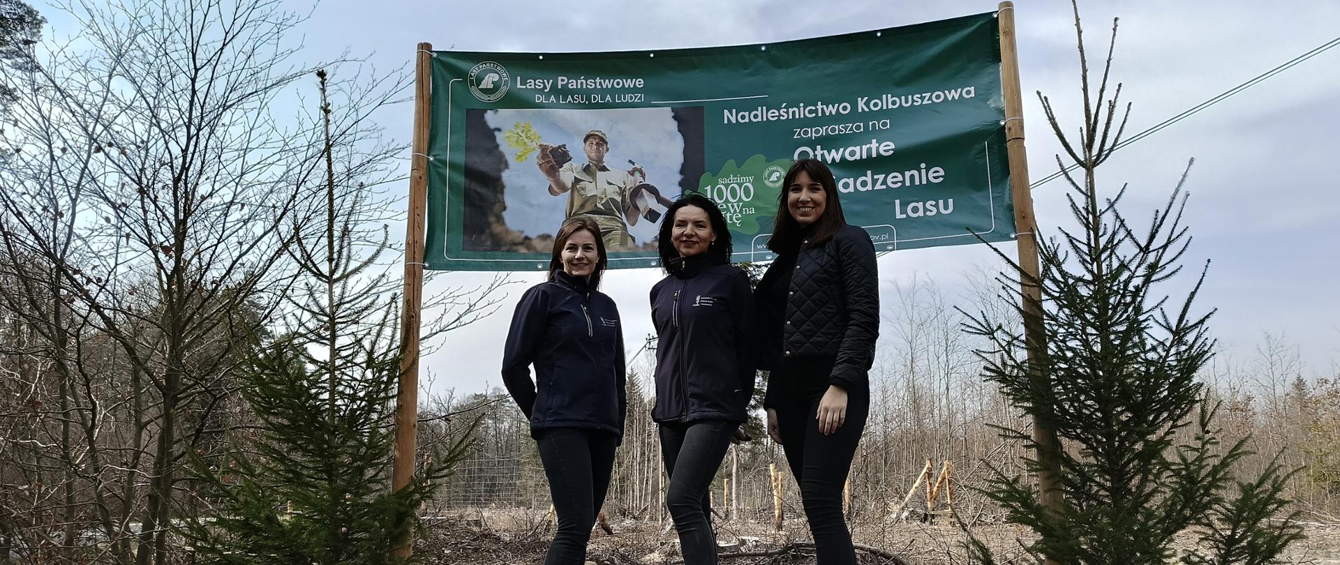 Trzy kobiety, ubrane na sportowo w ciemne ubrania. Stoją przed dużym zielonym banerem. W tle widać las. Obok banneru rosną dwa średniej wielkości drzewka. Przed kobietami na ziemi widać pień z uciętego wcześniej drzewa. 