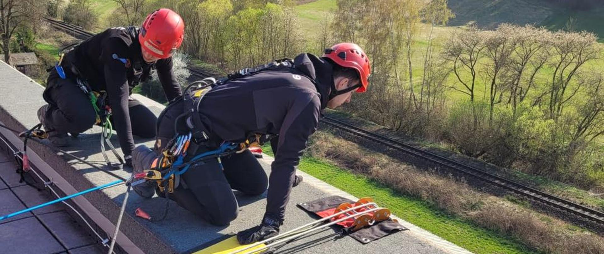 Ratownicy wysokościowi przygotowują stanowisko linowe na krawędzi dachu podczas ćwiczeń z technik dostępu linowego. W tle rozciąga się malowniczy krajobraz zielonych łąk i lasów