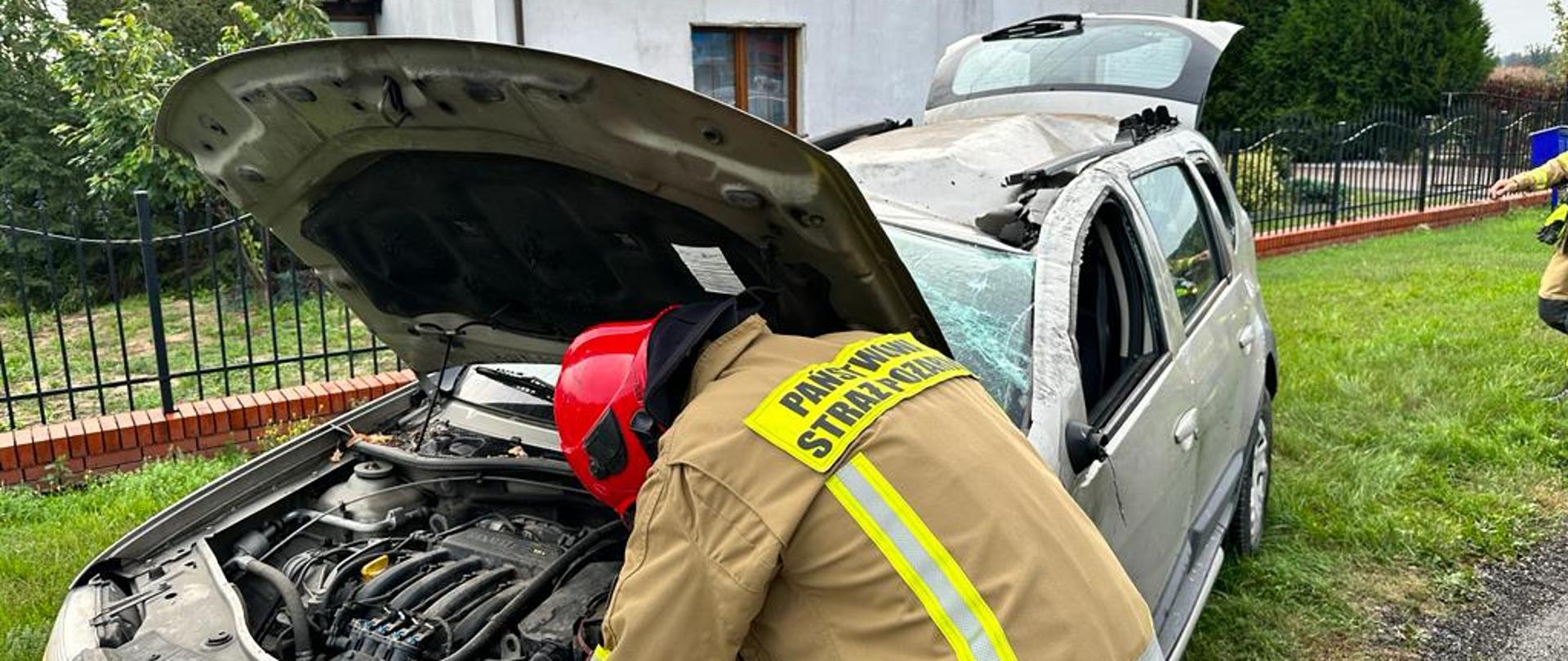 Samochód osobowy po dachowaniu stoi na kołach z podniesioną maską i klapą bagażnika. Strażak odłącza klemy akumulatora. W tle budynek mieszkalny.