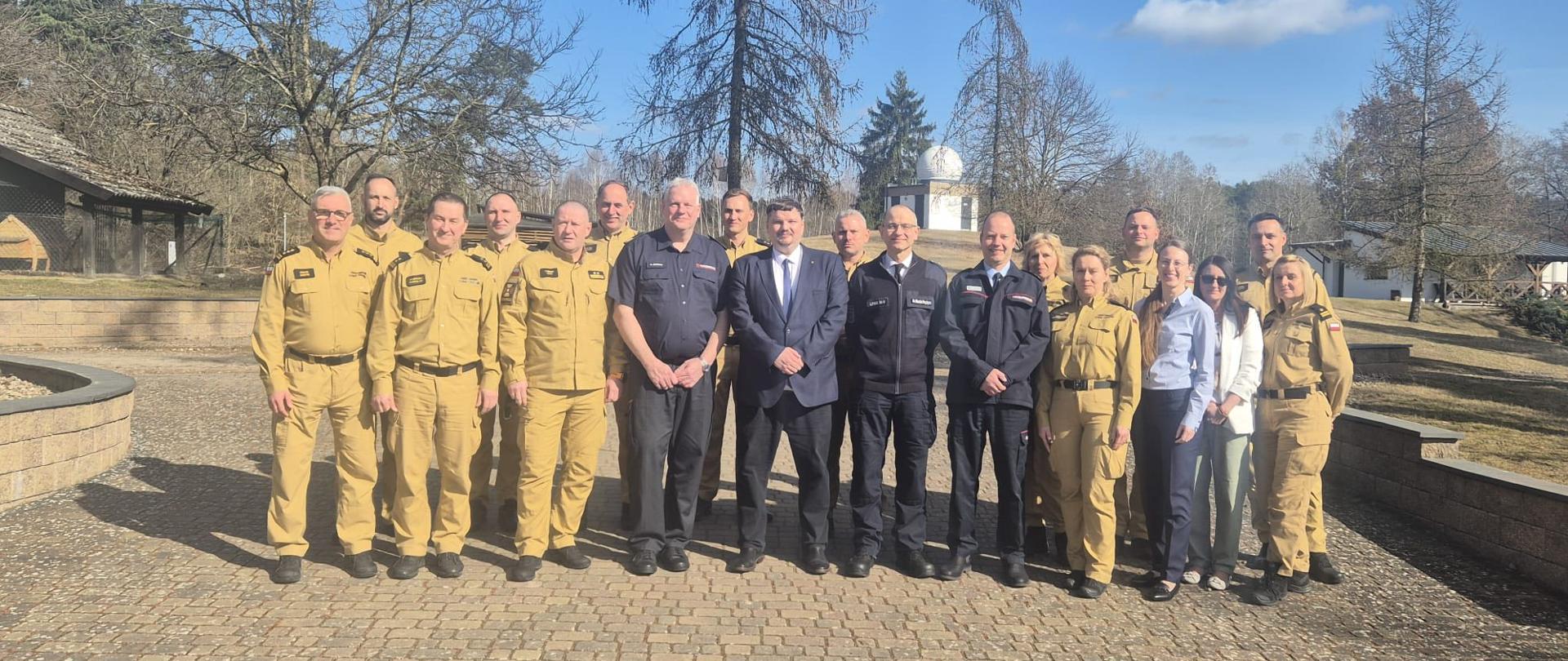 Duża grupa osób stoi na zewnątrz na brukowanym placu w słoneczny dzień. Wśród nich są strażacy w mundurach oraz osoby w ubraniach cywilnych. Wszyscy ustawieni są w jednym rzędzie do wspólnego zdjęcia, a w tle widać drzewa i budynki.