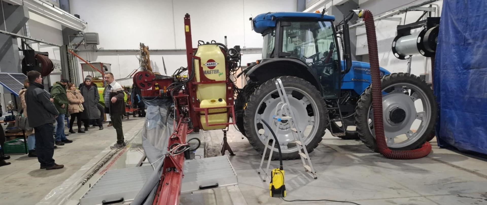 Zdjęcie przedstawia dużą halę. W jej centralnej części stoi ciągnik wraz z przymocowanym do niego opryskiwaczem polowym. Przy jednej ze ścian stoi grupa osób wraz z wykładowcą. 