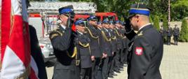 Zdjęcie zrobione na zewnątrz, podczas uroczystych obchodów Dnia Strażaka. Zdjęcie zrobione na placu przed jednostką ratowniczo-gaśniczą. Oficer Państwowej Straży Pożarnej w stopniu starszego brygadiera oddaje salut strażakowi. Obok strażaka stoją uczestnicy uroczystości - strażacy Państwowej Straży Pożarnej - wszyscy ubrani są w mundury galowe. Za nimi ustawione są samochody strażackie.