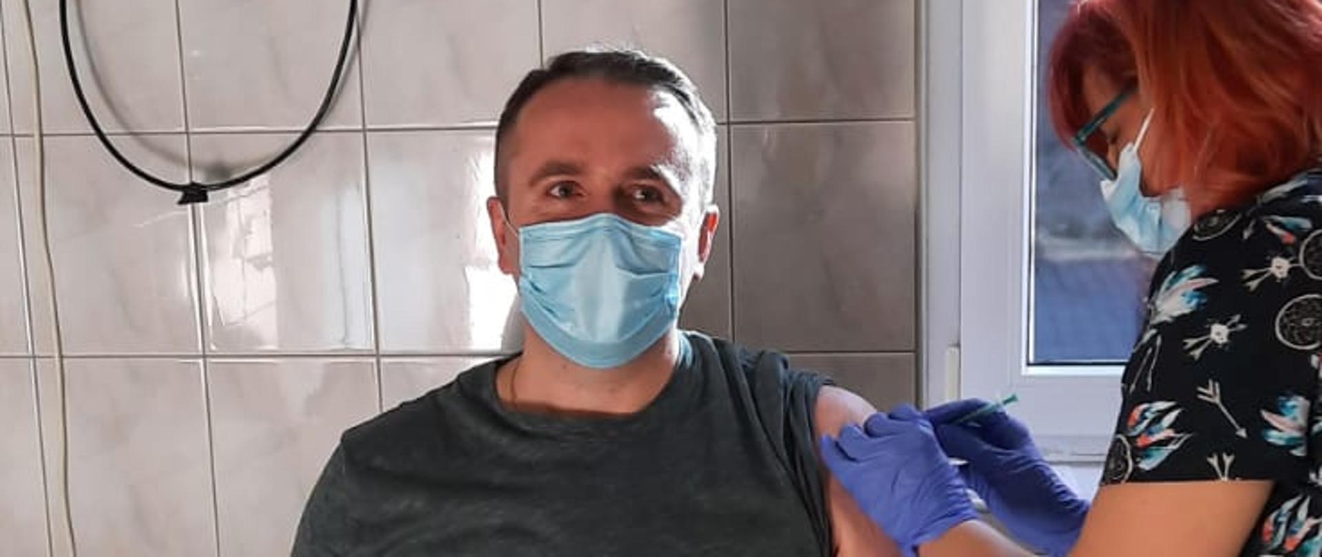 Szczepienia odbyły się przed południem w szpitalu w Gryfinie, w gabinecie zabiegowym, strażak siedzi na krześle z jego lewej strony Pani pielęgniarka podająca szczepionkę w lewe ramię, oboje są w maseczkach, pielęgniarka dodatkowo w rękawiczkach lateksowych. Na zdjęciu znajduje się mł. bryg. Krystian Kowal - zastępca komendanta powiatowego psp w Choszcznie