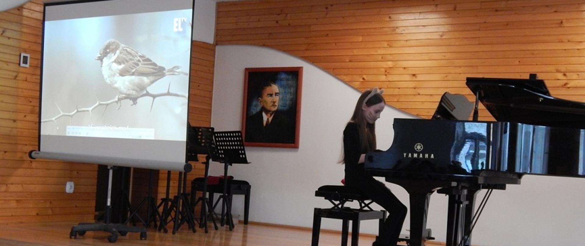 dziewczynka grająca na czarnym fortepianie, w tle ekran przedstawiający sylwetkę wróbla