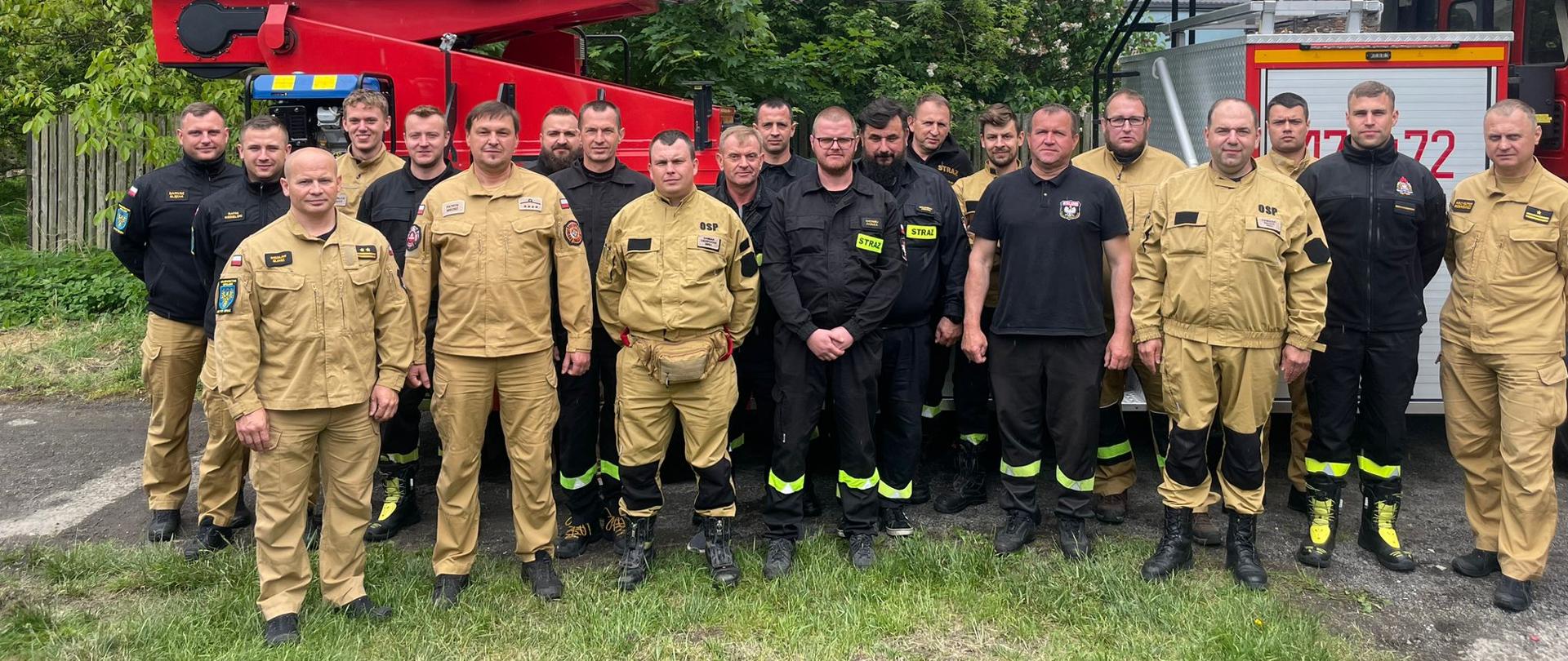 Strażacy Państwowej Straży Pożarnej oraz druhowie jednostek OSP w umundurowaniu służbowym i koszarowym stoją na tle drabiny mechanicznej do wspólnej fotografii.