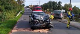 Dwa rozbite auta stoją na drodze, wśród nich strażacy usuwający skutki wypadku