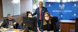 Na zdjęciu wojewoda opolski z pracownikami urzędu, siedzącymi za biurkiem w punkcie recepcyjnym w Opolu. W tle baner Wojewoda Opolski. 