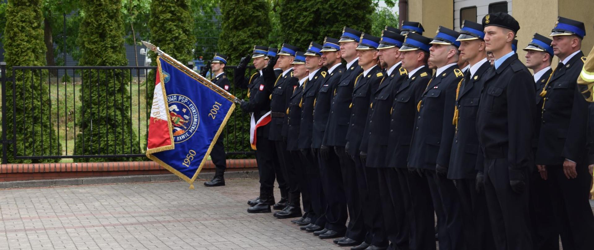 Pododdział stoi w strojach galowych w dwurzędzie. Na początku jeden z strażaków trzyma sztandar komendy skierowany do przodu. 
