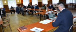 Komendant Powiatowy siedzi za stołem. przed nim leży laptop i notatki i rzutnik multimedialny. na dalszym planie siedzą za stołami strażacy.