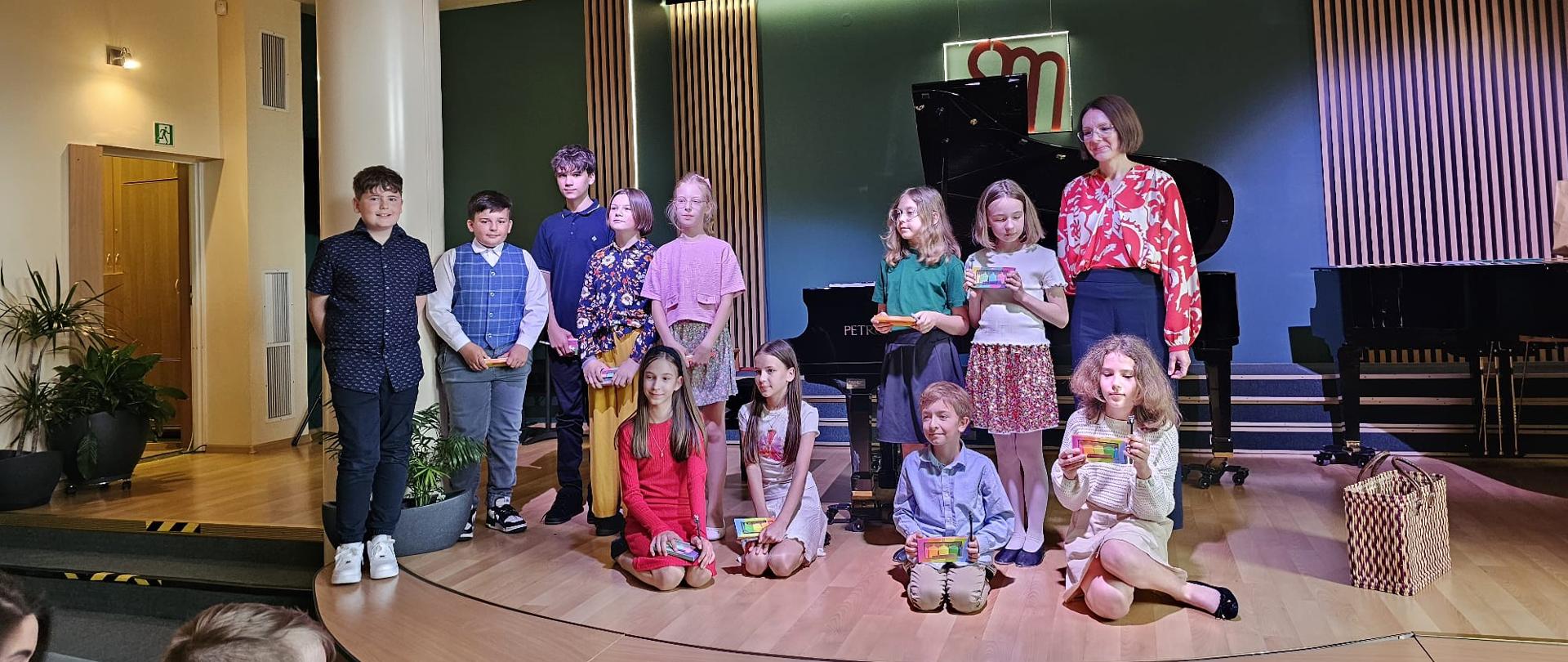 Na zdjęciu widać grupę dzieci oraz dorosłą osobę stojących na scenie. Wszyscy mają uśmiechnięte twarze, a niektóre dzieci trzymają w rękach kolorowe przedmioty, prawdopodobnie upominki lub dekoracje. Dorosła osoba, która stoi po prawej, jest ubrana w kolorową bluzkę i ciemną spódnicę. W tle znajduje się fortepian oraz dekoracje, a scena jest oświetlona. Z przodu zdjęcia widoczne są fragmenty krzeseł, co sugeruje, że publiczność siedziała przed sceną. Atmosfera wydaje się radosna i celebracyjna.
