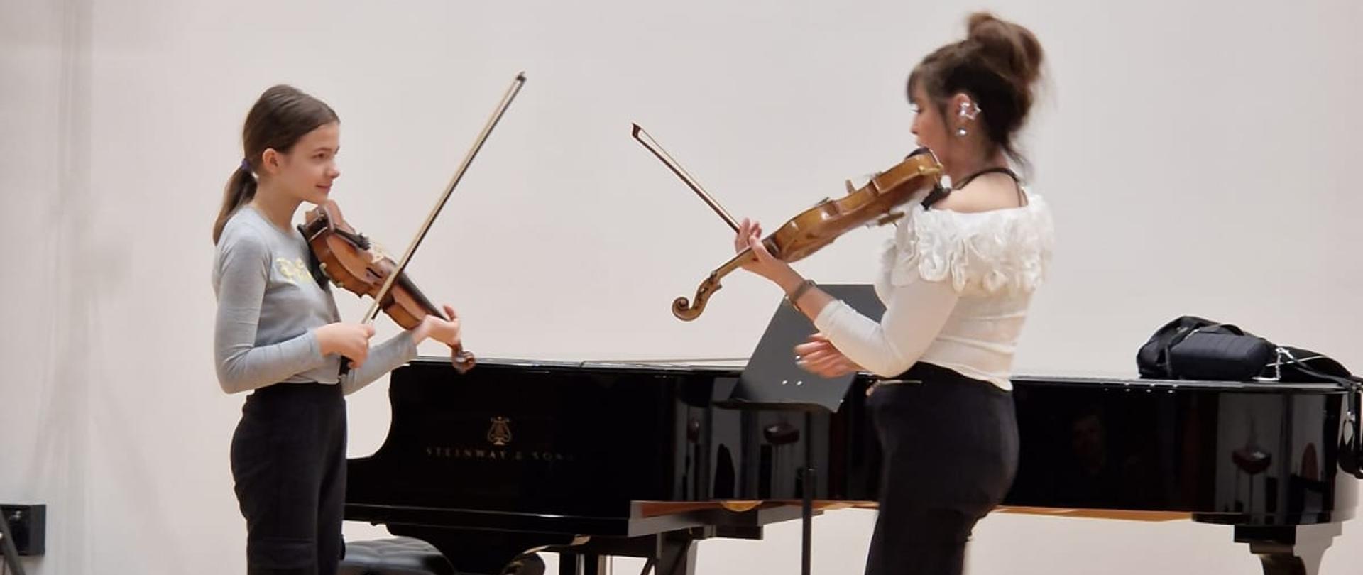 Na scenie z lewej Aleksandra Bartoszczyk - uczennica klasy skrzypiec p. Kingi Hudela - ze skrzypcami. z prawej prof. dr hab. Karina Gudaszewska ze skrzypcami (w trakcie lekcji otwartej). W tle fortepian. Z przodu sceny kompozycje ze sztucznych kwiatów (dekoracja w różnych odcieniach zieleni oraz kolorze białym i pudrowego różu).