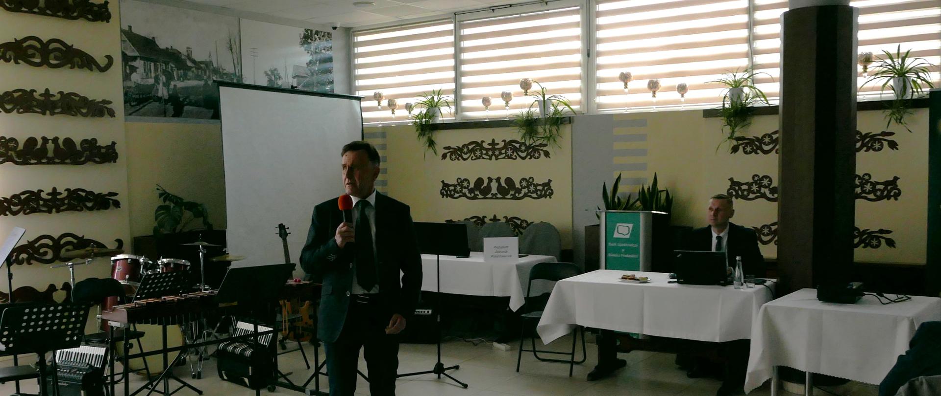 Pan z mikrofonem wita wszystkich przybyłych na zebranie i koncert. Za nim rozstawione pulpity i instrumenty muzyczne. Obok za stołem przy komputerze siedzi inny pan i słucha prowadzaącego.