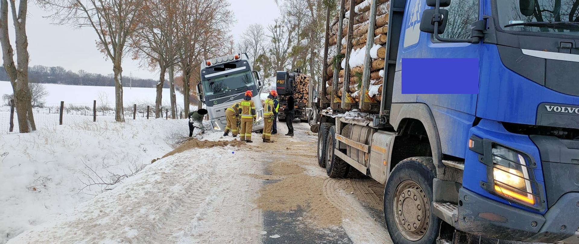 Zima, dużo śniegu. Na zdjęciu biała ciężarówka częściowo w rowie z prawej strony stoi niebieska ciężarówka z drzewem.